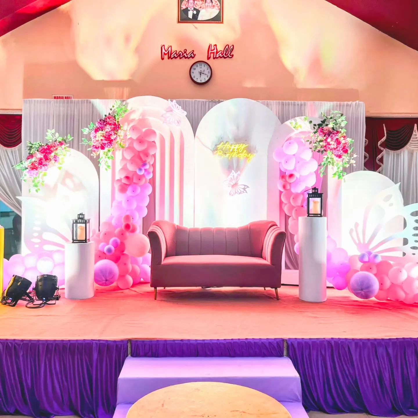 Pink and lavender butterfly-themed birthday stage with balloon arches and Happy Birthday neon